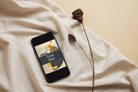 A smartphone lying on draped cream fabric, displaying a graphic titled 'Trail Unit' with a map and yellow route illustration. A dried flower stem rests beside the phone against a warm beige background.