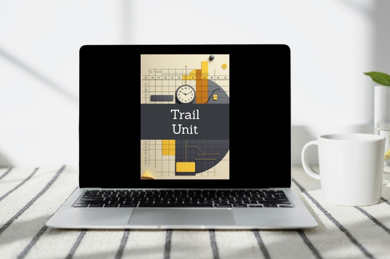 A laptop on a striped fabric surface with a white mug beside it. The laptop screen displays a book or cover design titled "Trail Unit" with a geometric illustration in beige, yellow, orange, and dark gray.